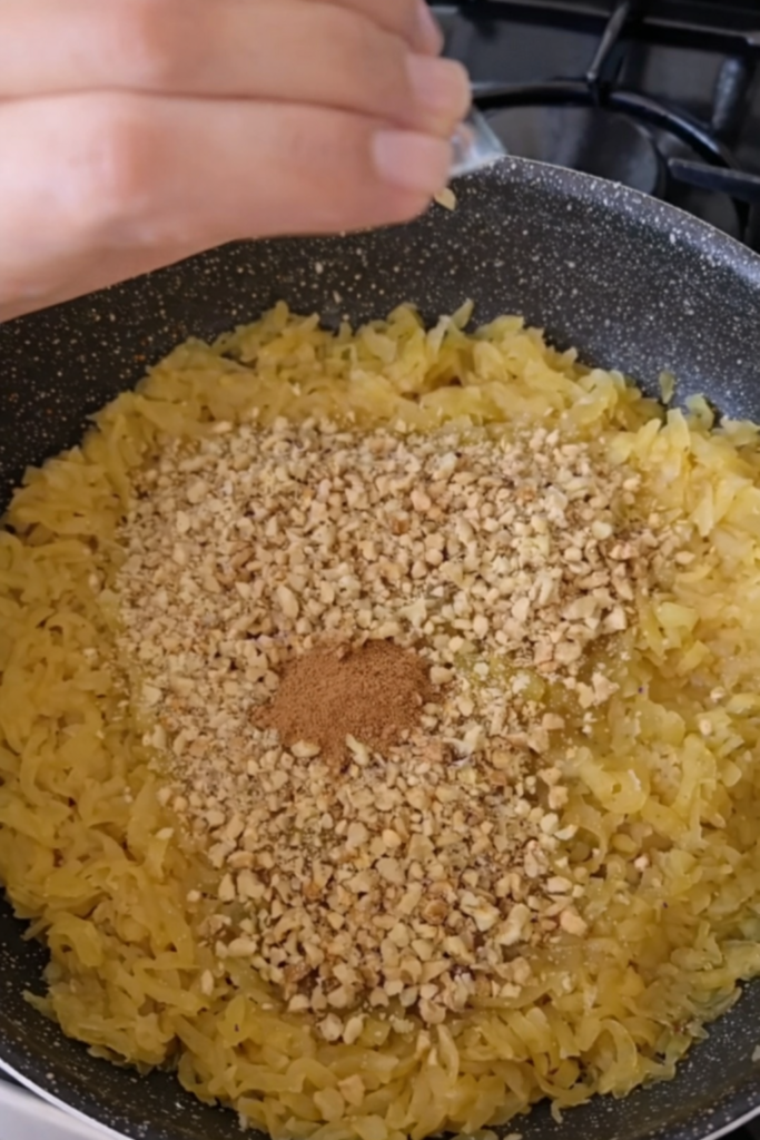 Na slici se vidi priprema fila od jabuka u tavi, gdje se narendane jabuke kuhaju zajedno sa mljevenim orasima i cimetom. U trenutku fotografije dodaje se cimet, dok su orasi ravnomjerno raspoređeni po površini smjese. Ovakav prikaz jasno pokazuje teksturu fila i redoslijed dodavanja sastojaka tokom pripreme domaćeg rolata od jabuka.