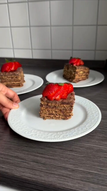 Komad čokoladnog kolača sa slojevima keksa i kreme, ukrašen jagodom na bijelom tanjiru