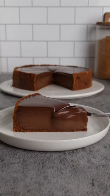 Kriška čokoladnog tarta s podlogom od keksa na bijelom tanjiru, s ostatkom tarta u pozadini