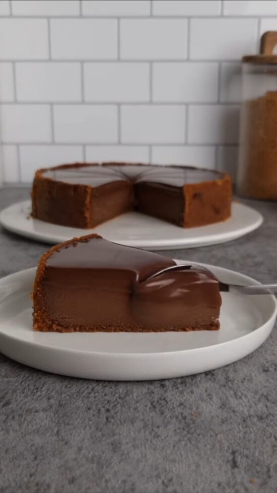 Kriška čokoladnog tarta s podlogom od keksa na bijelom tanjiru, s ostatkom tarta u pozadini